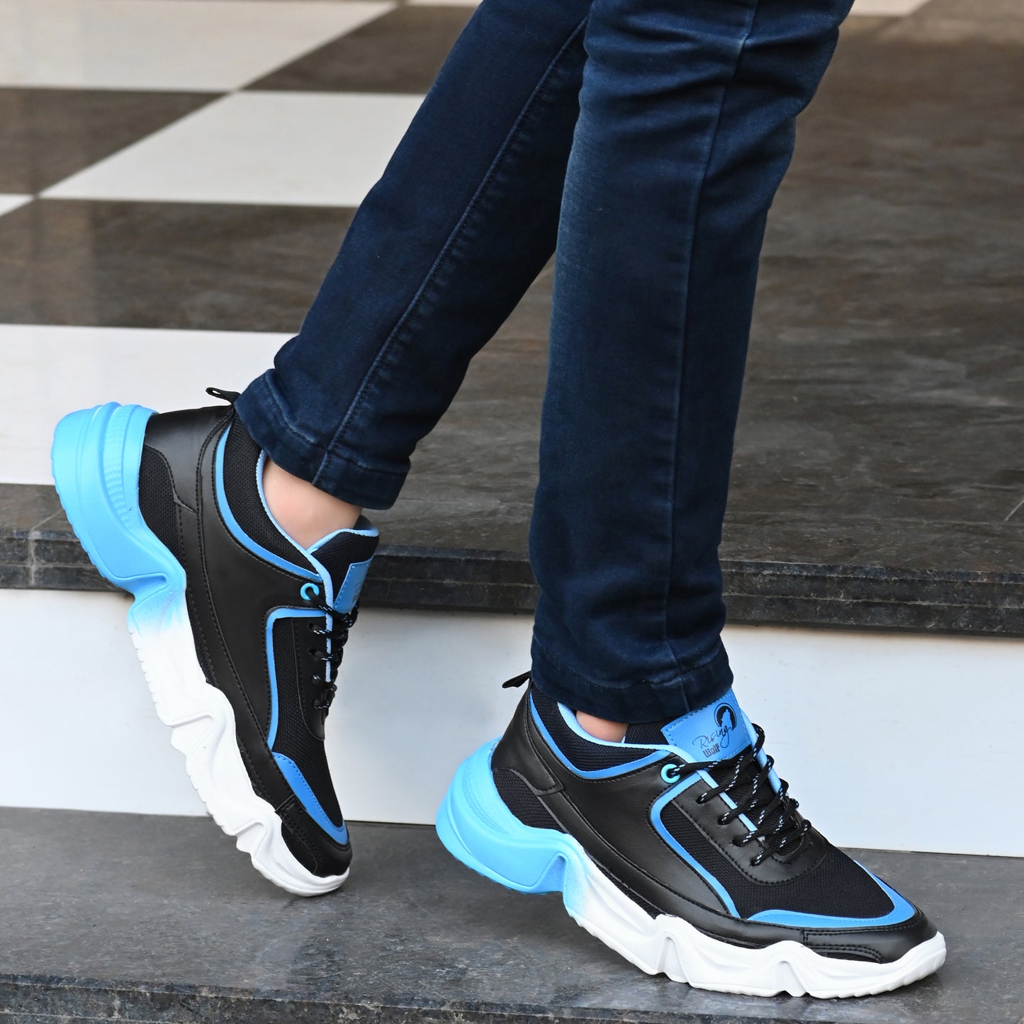 men's blue and black chunky sneakers with a white wavy sole, styled with dark blue denim jeans on a tiled staircase.