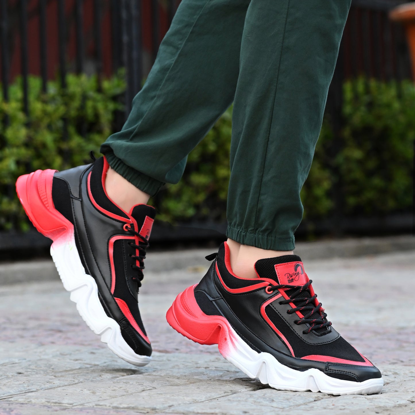 Rising Wolf men's chunky sneakers in black and red with a thick white wavy sole, styled with green joggers on a paved walk.